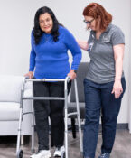 Photo of nurse in a gray polo and jeans helping a patient with a walker